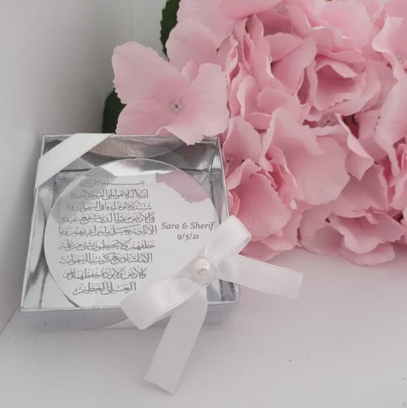 Op de afbeelding: Een zilveren doosje met een wit lint en een ronde, doorzichtige schijf erin. De schijf heeft Arabische kalligrafie en de tekst "Sara & Sherif 9/5/21".