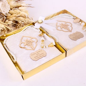 May include: Two gift boxes with cream-colored prayer mats, adorned with gold accents and pearl-like beads. Each box contains a prayer mat, a wooden ornament, and a string of beads. The boxes are tied with a white ribbon and a decorative flower.