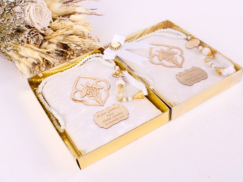 May include: Two gift boxes with cream-colored prayer mats, adorned with gold accents and pearl-like beads. Each box contains a prayer mat, a wooden ornament, and a string of beads. The boxes are tied with a white ribbon and a decorative flower.