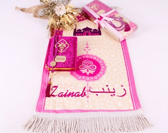 Peut inclure: Un tapis de prière rose avec une frange blanche, un livre de prière en velours rose avec une inscription dorée qui dit "Zainab" et un collier de prière en perles roses. Le tapis a un motif géométrique rose et blanc et le nom "Zainab" écrit en arabe.