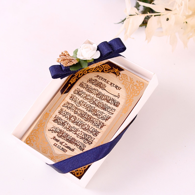 May include: A white box with a blue ribbon and a white flower on top. Inside the box is a wooden plaque with gold lettering that says "Ayatul Kursi" and a date. The plaque is in a gold frame.