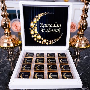 May include: A white box of chocolates, each with a crescent moon and star design. The box lid features a crescent moon made of hanging lanterns, with the words "Ramadan Mubarak" in white script. Gold candle holders are on either side.