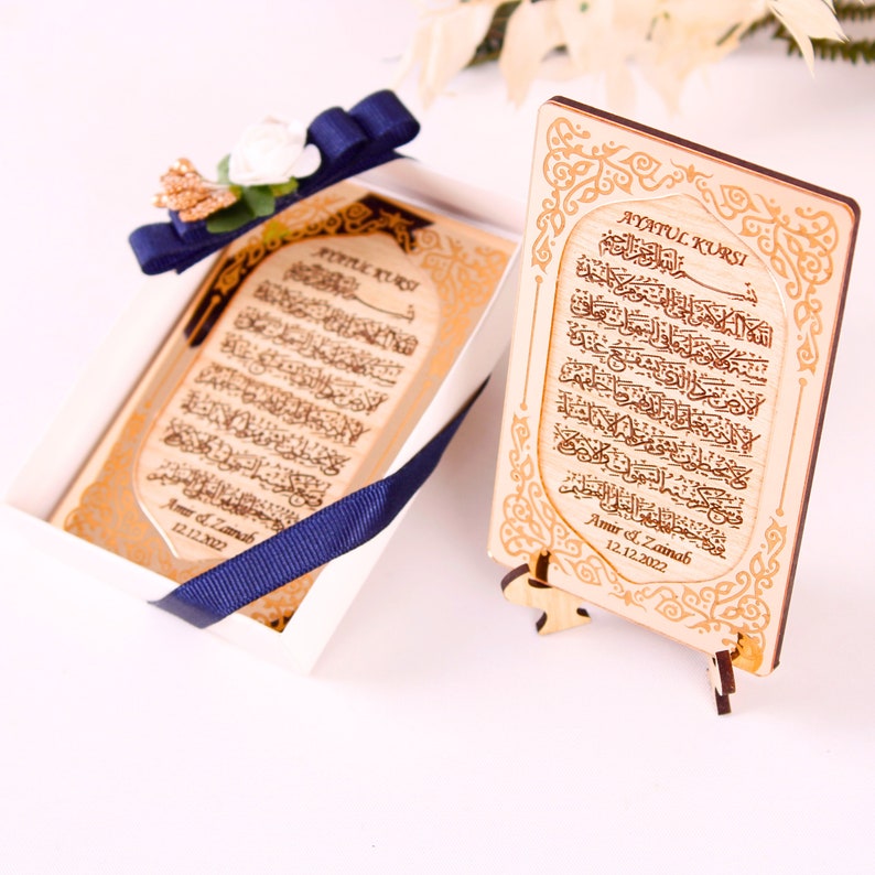 May include: A wooden plaque with a gold-colored design and Arabic calligraphy. The plaque is engraved with the text "Ayatul Kursi" and the names "Amir & Zainab" with the date "12.12.2022". The plaque is on a small wooden stand.