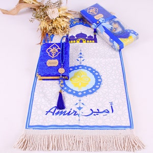 May include: A blue prayer rug with a white background and a blue and yellow design. The rug has a fringe border and the name "Amir" is embroidered on it. The rug is accompanied by a blue velvet prayer book with the name "Amir" on it and a blue prayer bead set.