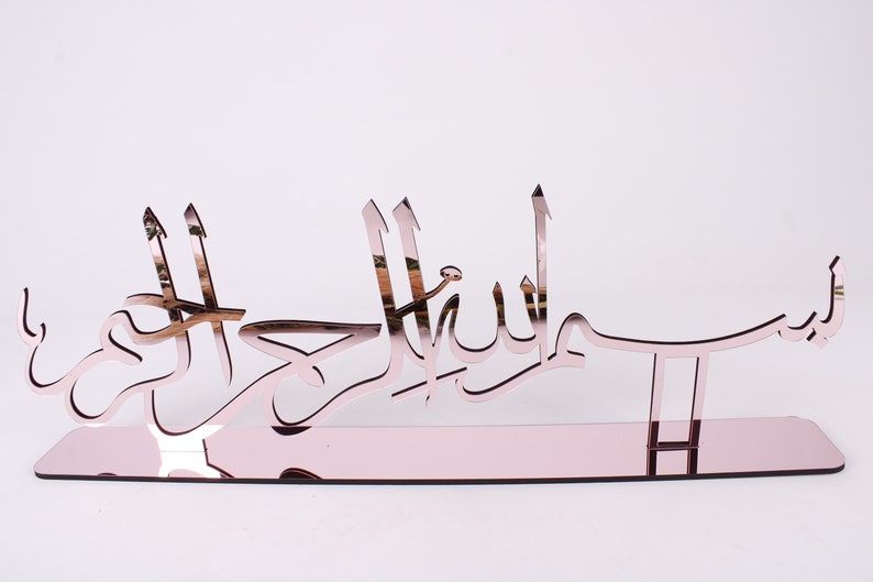 May include: A rose gold mirrored wall decor with the Arabic phrase "Bismillah" in a decorative font.