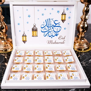 May include: A white gift box with a blue and gold design featuring a traditional Islamic greeting, "Eid Mubarak", and hanging lanterns. The box is filled with 36 small, square chocolates, each with a similar design.