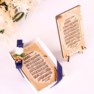 May include: A gold-colored wooden plaque with Arabic calligraphy and the text "Ayatul Kursi" in English. The plaque is in a white box with a blue ribbon and a white flower. The plaque is on a small wooden stand.