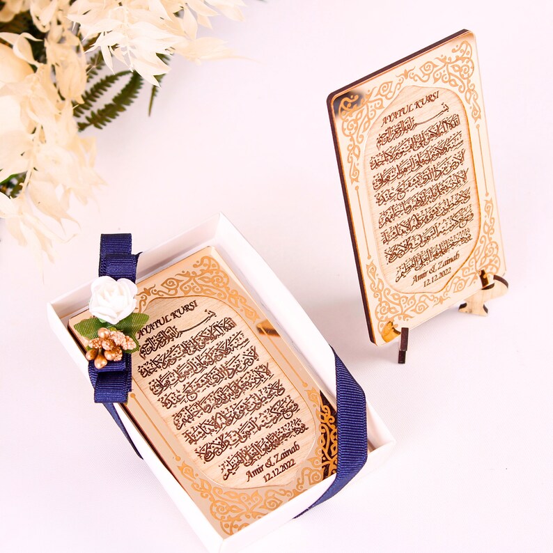 May include: A gold-colored wooden plaque with Arabic calligraphy and the text "Ayatul Kursi" in English. The plaque is in a white box with a blue ribbon and a white flower. The plaque is on a small wooden stand.