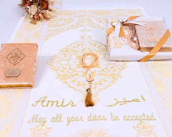 May include: A white prayer rug with gold embroidery and the name "Amir" embroidered on it. The rug is part of a gift set that includes a prayer book, prayer beads, and a small decorative box. The gift set is wrapped in a white box with a gold ribbon.