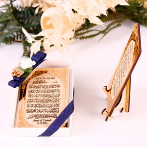 May include: A wooden plaque with gold Arabic calligraphy and the text "Ayatul Kursi!"  The plaque is in a white box with a blue ribbon and a small white flower. The plaque is also on a wooden stand.