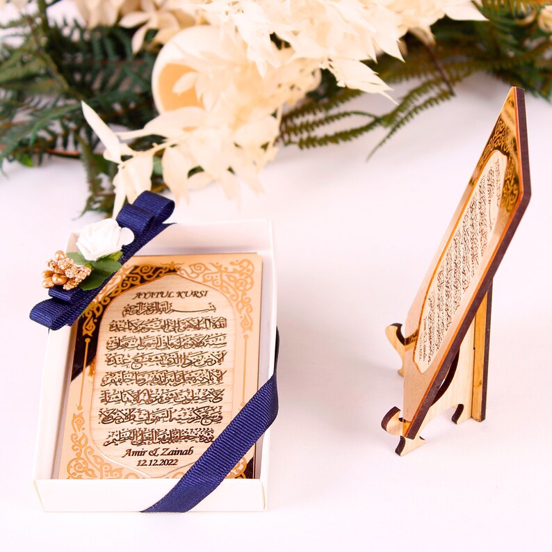 May include: A wooden plaque with gold Arabic calligraphy and the text "Ayatul Kursi!"  The plaque is in a white box with a blue ribbon and a small white flower. The plaque is also on a wooden stand.