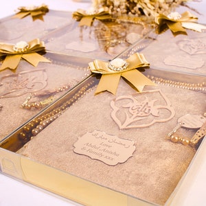 May include: Gold gift boxes with clear lids containing a gold prayer rug, a set of prayer beads, and a wooden ornament with Arabic calligraphy. The boxes are tied with gold ribbon and have a personalized tag that reads "Ramadan Mubarak Love Abdul, Anisha & Family xxx".