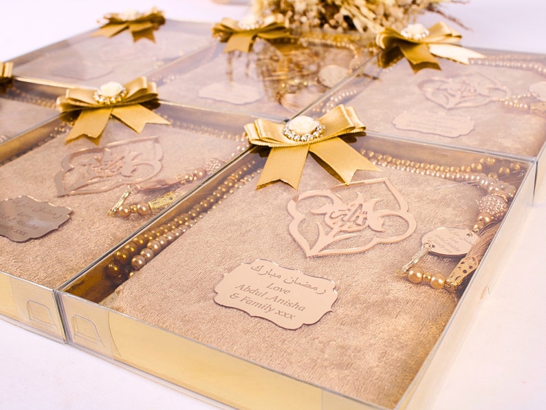 May include: Gold gift boxes with clear lids containing a gold prayer rug, a set of prayer beads, and a wooden ornament with Arabic calligraphy. The boxes are tied with gold ribbon and have a personalized tag that reads "Ramadan Mubarak Love Abdul, Anisha & Family xxx".