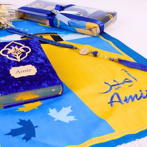May include: A blue prayer mat with a yellow central panel, displaying the name "Amir" in Arabic and English. A blue velvet book with "Amir" on the cover and a blue beaded rosary are also visible. A gift box tied with blue ribbon is in the background.