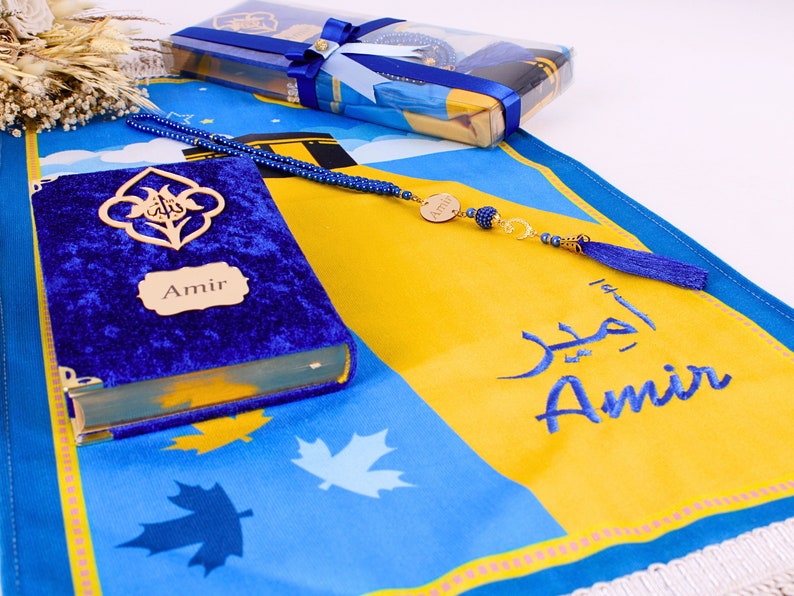 May include: A blue prayer mat with a yellow central panel, displaying the name "Amir" in Arabic and English. A blue velvet book with "Amir" on the cover and a blue beaded rosary are also visible. A gift box tied with blue ribbon is in the background.
