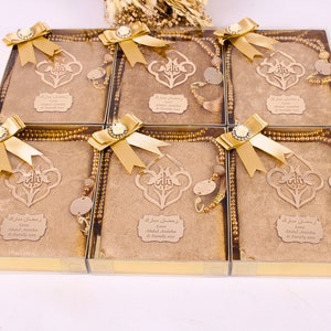 May include: Six gold gift boxes with clear lids, each containing a brown prayer rug, a gold prayer bead necklace, and a wooden plaque with Arabic calligraphy. The boxes are decorated with gold bows and a white flower.