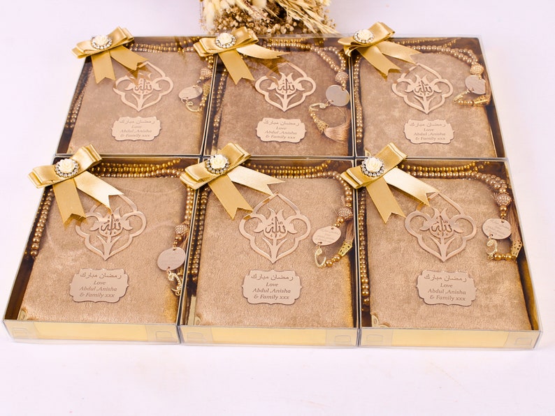 May include: Six gold gift boxes with clear lids, each containing a brown prayer rug, a gold prayer bead necklace, and a wooden plaque with Arabic calligraphy. The boxes are decorated with gold bows and a white flower.