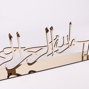 May include: Gold-colored metal wall decor with Arabic calligraphy that reads "Subhanallah" in a modern style. The decor is mounted on a mirrored stand.