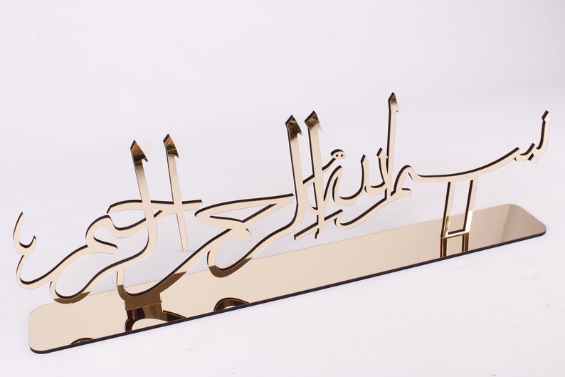 May include: Gold-colored metal wall decor with Arabic calligraphy that reads "Subhanallah" in a modern style. The decor is mounted on a mirrored stand.