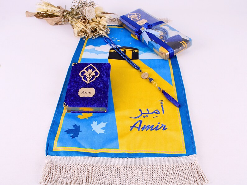 May include: A blue and yellow prayer mat with the name "Amir" embroidered in blue. A blue velvet Quran with gold accents and a matching prayer bead set are included. A gift set with a floral arrangement.