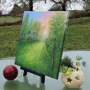 Puede incluir: Pintura de paisaje original sobre lienzo, que representa un camino soleado a trav&eacute;s de un bosque verde. La obra de arte se exhibe en un peque&ntilde;o caballete negro. Tambi&eacute;n hay un adorno rojo brillante y un portavelas con forma de estrella.