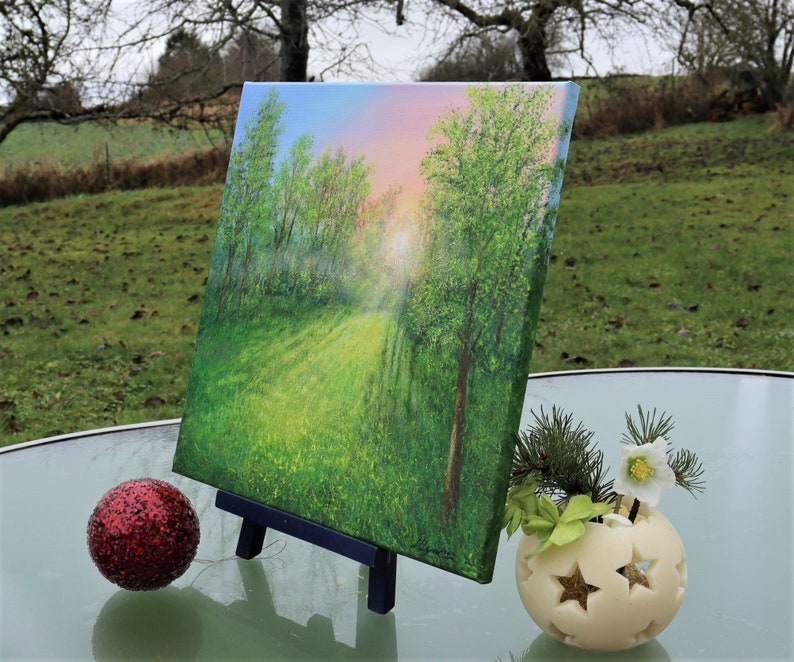 Puede incluir: Pintura de paisaje original sobre lienzo, que representa un camino soleado a trav&eacute;s de un bosque verde. La obra de arte se exhibe en un peque&ntilde;o caballete negro. Tambi&eacute;n hay un adorno rojo brillante y un portavelas con forma de estrella.