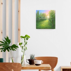 Puede incluir: Cuadro cuadrado que representa una escena de bosque soleado. La pintura muestra un camino que conduce a la distancia, rodeado de &aacute;rboles verdes y un campo de flores amarillas. El cielo pasa del azul al rosa, sugiriendo el amanecer o el atardecer.