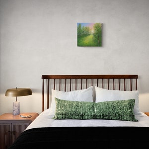 Puede incluir: Una pintura cuadrada que representa un paisaje con un camino que conduce a un bosque. La obra de arte est&aacute; colgada sobre una cama con ropa de cama blanca, un coj&iacute;n verde y una manta negra. Dos l&aacute;mparas con bases de m&aacute;rmol flanquean la cama.
