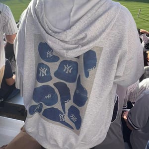 May include: A light gray hoodie with a blue graphic of New York Yankees baseball caps. The caps are in various shapes and orientations. The hoodie has a hood and is being worn at a baseball game.