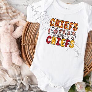 May include: White baby bodysuit with the word "CHIEFS" in red, yellow, and white block letters. The bodysuit is on a woven basket with a stuffed animal and a patterned cloth.