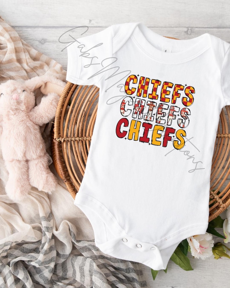 May include: White baby bodysuit with the word "CHIEFS" in red, yellow, and white block letters. The bodysuit is on a woven basket with a stuffed animal and a patterned cloth.