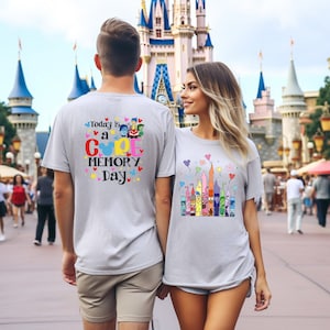 May include: Two light grey t-shirts with colourful graphics and text. One shirt says "Today is a COOL MEMORY Day" with cartoon characters. The other shirt features a castle design with cartoon characters and fireworks.