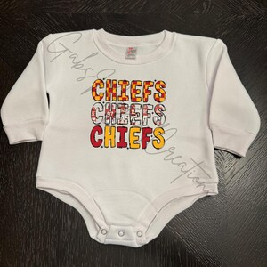May include: White baby bodysuit with long sleeves. The front features the word "CHIEFS" in a stacked design, with each word in a different color and pattern, including red, yellow, and black. The bodysuit has snap closures.