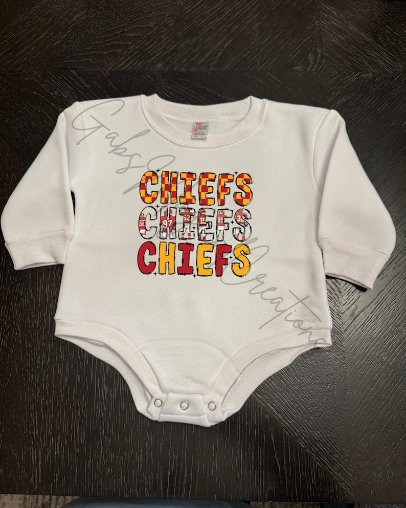 May include: White baby bodysuit with long sleeves. The front features the word "CHIEFS" in a stacked design, with each word in a different color and pattern, including red, yellow, and black. The bodysuit has snap closures.