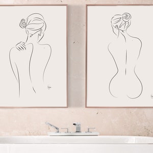 Puede incluir: Dos dibujos a línea enmarcados de una figura desnuda en un estilo minimalista. Las figuras están de espaldas al espectador y tienen el pelo recogido en un moño. Los dibujos están colgados en una pared rosa claro sobre una bañera blanca.