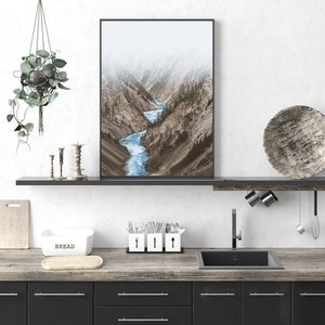 May include: A framed photograph of a river winding through a mountain valley. The river is a bright blue and the mountains are a muted brown. The photograph is hanging on a white wall above a kitchen counter.