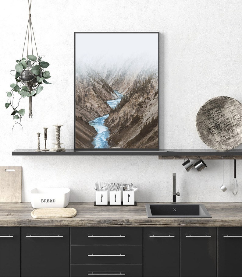 May include: A framed photograph of a river winding through a mountain valley. The river is a bright blue and the mountains are a muted brown. The photograph is hanging on a white wall above a kitchen counter.