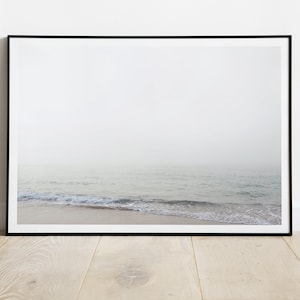 May include: A framed print featuring a serene coastal scene. The photograph captures a misty ocean horizon with gentle waves lapping a sandy shore. The print is encased in a sleek black frame, adding a modern touch to the minimalist aesthetic.