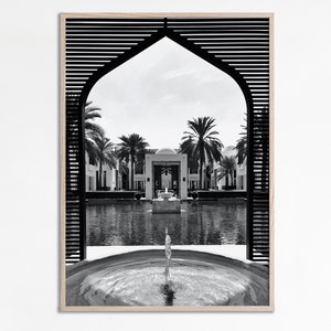May include: Black and white photograph of a fountain in a courtyard with palm trees and a building in the background. The fountain is framed by a wooden archway with a lattice design.