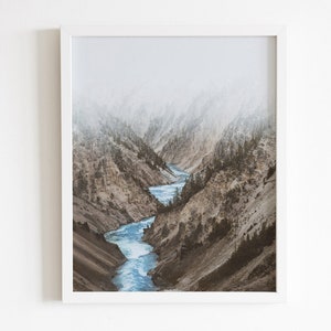 May include: A misty mountain landscape with a winding river flowing through a canyon. The river is a bright blue, and the mountains are a muted brown and green.