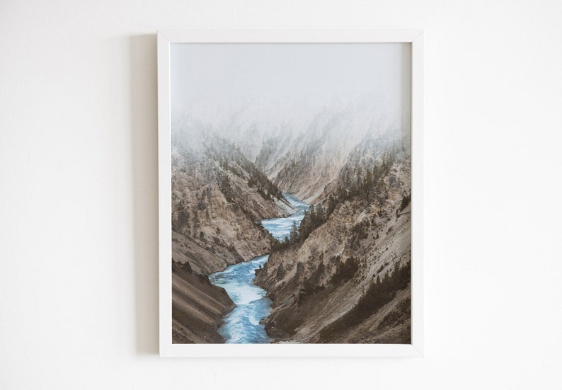 May include: A misty mountain landscape with a winding river flowing through a canyon. The river is a bright blue, and the mountains are a muted brown and green.