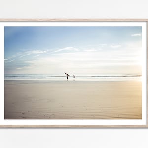 Op de afbeelding: Twee mensen lopen langs een zandstrand met surfplanken in de hand, de oceaan achter hen en een blauwe lucht erboven.