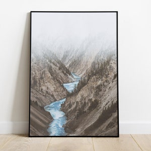 May include: A misty mountain landscape with a winding river flowing through a canyon. The river is a bright blue, and the mountains are a muted brown and green.