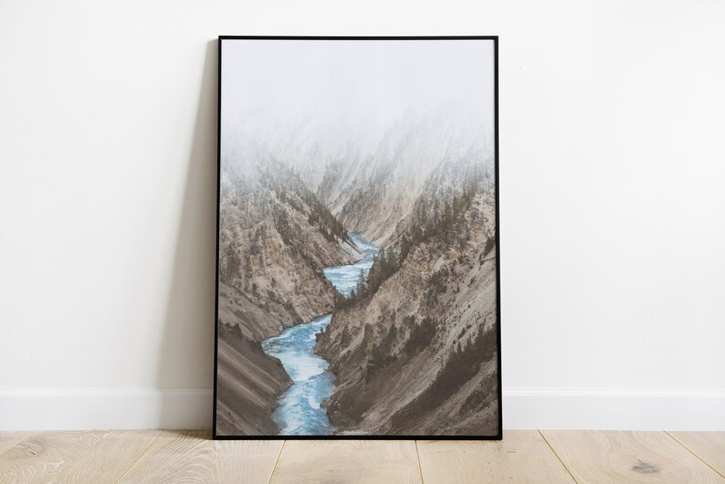 May include: A misty mountain landscape with a winding river flowing through a canyon. The river is a bright blue, and the mountains are a muted brown and green.