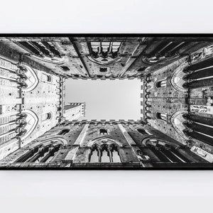 May include: Black and white photograph of a building with a square opening in the center, looking up towards the sky. The building has a textured stone exterior and arched windows.