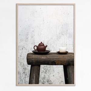 Può includere: Fotografia incorniciata con una teiera marrone e una tazza bianca, ciascuna su un piattino, appoggiate su uno sgabello di legno rustico. Lo sfondo è un muro bianco strutturato, minimalista.