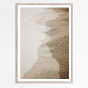 Puede incluir: Una fotografía enmarcada de una escena de playa con un primer plano de la arena y el agua. La arena es de color marrón claro y el agua es de color azul claro. El agua se está retirando de la costa, creando un patrón ondulado en la arena.