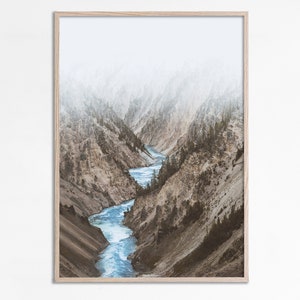 May include: A photograph of a river winding through a canyon in a mountainous landscape. The river is a bright blue and the mountains are a muted brown and gray. The sky is a hazy white.