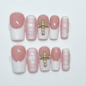 May include: A set of press-on nails featuring a French manicure design with a light pink and white color scheme. Some nails have a gold cross with pearl accents. The nails are displayed on a white surface.