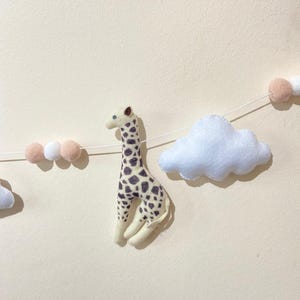 May include: A string of felt decorations featuring a giraffe with brown spots, white pom-poms, and a white cloud. The giraffe is a decorative item, possibly for a nursery or children's room.